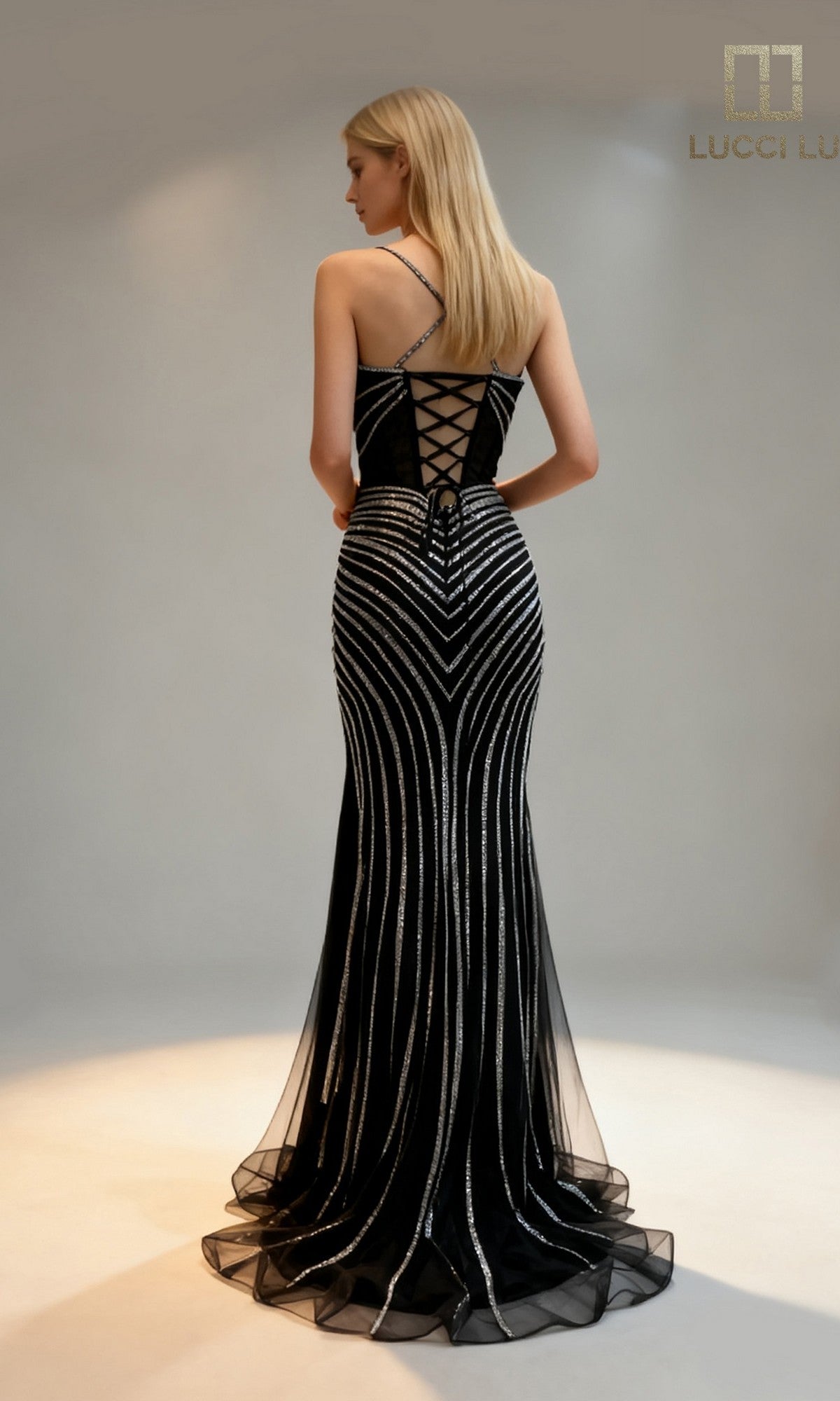 A woman faces away wearing the black Lucci Lu 1448 long prom dress, featuring silver vertical and diagonal stripes, a lace-up corset back, and thin straps.